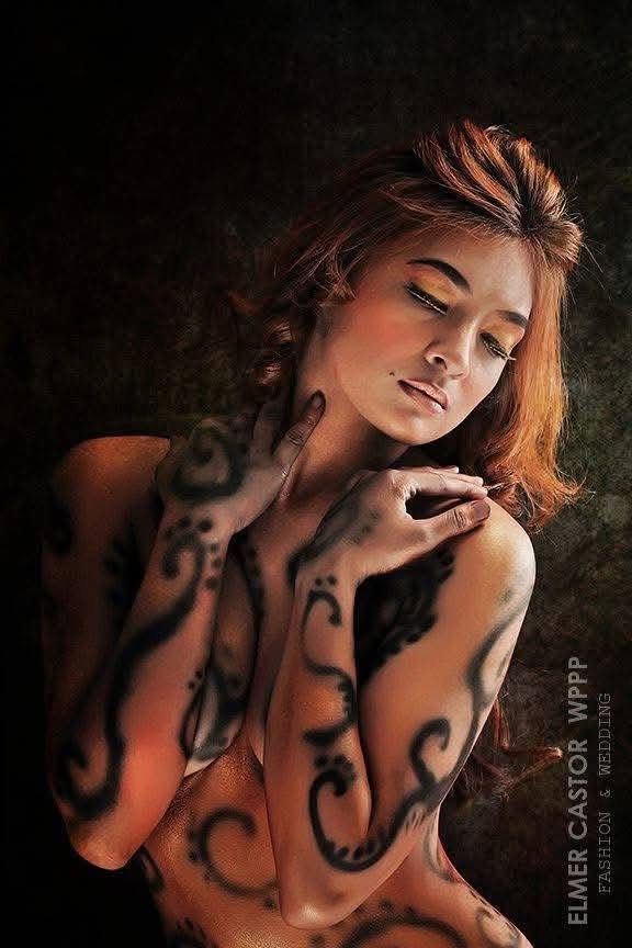 Model with intricate black swirl body paint across her shoulders, neck, and arms, softly lit against a dark background.